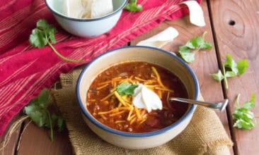 A blue rimmed, white bowl sits on a burlap napkin a top a wood slat table. The bowl is full of the perfect chili, topped with shredded cheese, sour cream and, garnished with cilantro.