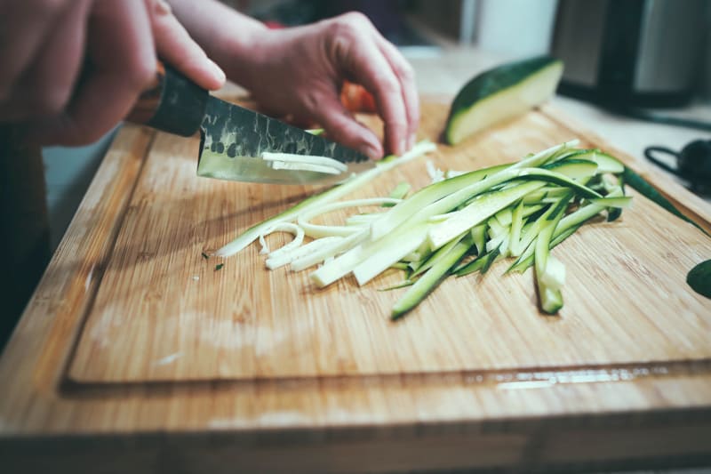 A butcher block on a counter top with a zucchini being sliced julienne by someone.