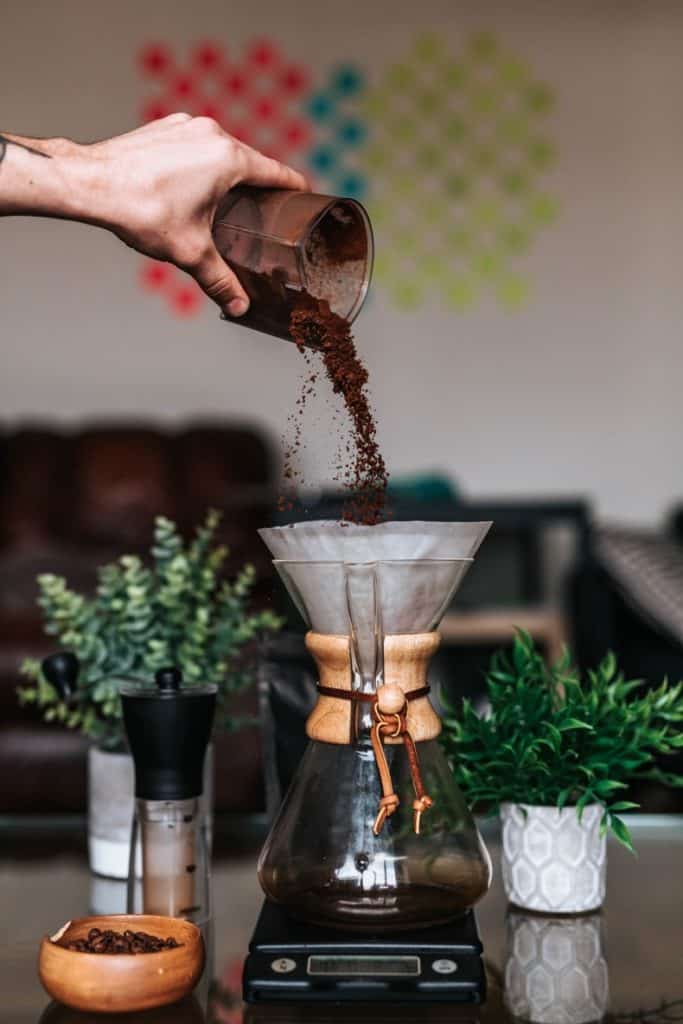 hand pouring coffee grounds into a chemex pourover coffee maker with decorative elements beside it to indicate gift ideas