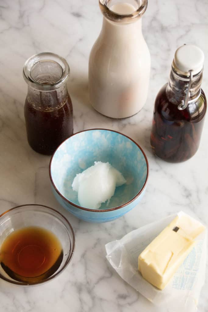 Ingredients for butter chai latte sit in glass bottles, small blue pottery bowl and a glass bowl on a marble counter.
