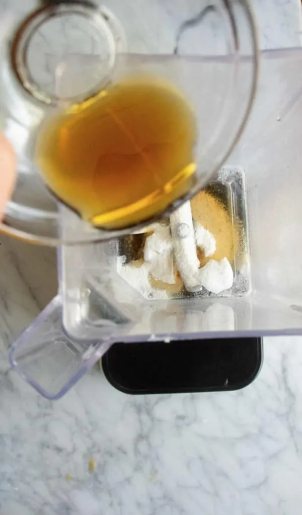 Bird's eye view of butter, maple syrup, coconut oil, vanilla extract, salt and spices in a blender with chai concentrate being poured into the blender.