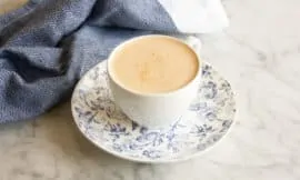 Butter Chai Latte in a white teacup sits on a blue and white saucer on a marble counter. A blue tea towel sits in the background.