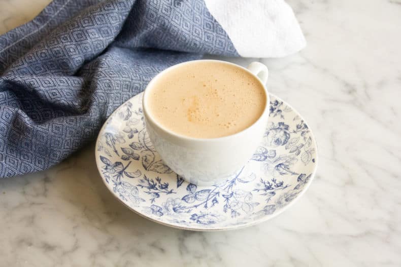 Butter Chai Latte in a white teacup sits on a blue and white saucer on a marble counter. A blue tea towel sits in the background. 