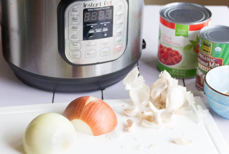 Close up shot of onions chopped on a white cutting board. Instant Pot and cans of diced tomatoes and coconut milk sit in the background.