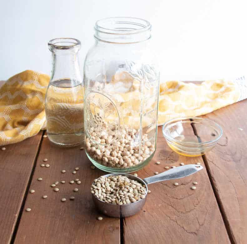 On top of a wooden table sits an open glass jar with lentils and chickpeas inside. A measuring cup sits infront full of legumes, and a small decanter of water is next to the jar.