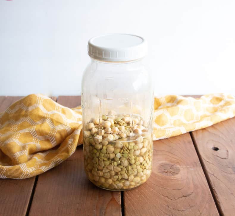 A jar of lentils and chickpeas that have been soaked sit on a wooden surface with a yellow tea towel nearby.