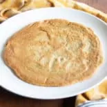 On top of a brown wood surface, a cooked lentil and chickpea flatbread sits on a white plate, a yellow tea towel is nearby.