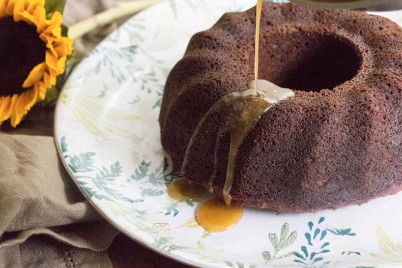 Gingerbread bundt cake sits on a white botanical plate as a golden maple glaze is poured over the cake.
