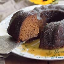 Maple Glazed Gingerbread Bundt cake sits on a white plate with a soft botanical pattern. A piece of cake has been cut and is being lifted with a silver cake slicer.