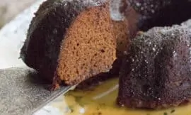 Maple Glazed Gingerbread Bundt cake sits on a white plate with a soft botanical pattern. A piece of cake has been cut and is being lifted with a silver cake slicer.