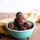 A teal bowl with a pyramid of chocolate macaroons sits on a brown wood table with a yellow tea towel deoratively placed in the background.