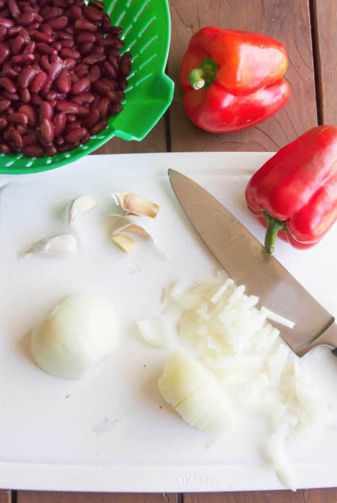 An onion is chopped on a white cutting board. A knife rests to the right. Garlic, two red peppers and beans in a green strainer sit nearby.