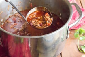 A full ladel lifts chili out of a large stainless steel pot full of the perfect cooked chili.