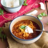 a bowl of chili in a white bowl, sitting on a wood surface, surrounded by a red linen, cilantro, and a bowl of sour cream