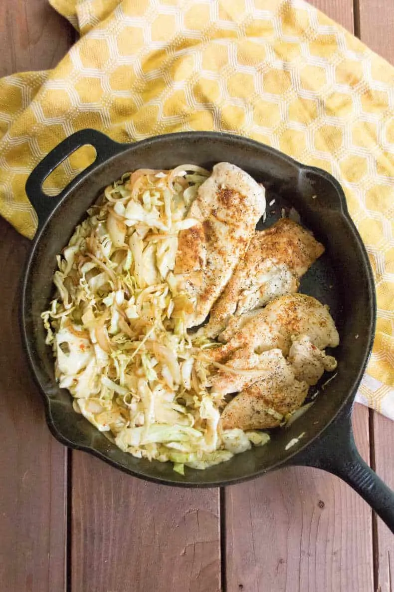 A view from above, a cast iron pan full of smoky cabbage noodles and cooked chicken breasts, sits on a yellow dish towel a top a wood slat table.