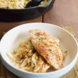 Resting on a burlpa npakin atop a wood slat tabletop is Seared chicken breast on top of a bed of smoky cabbage noodles in a white bowl. In the background sit the cast iron pan with chicken and cabbage noodles.