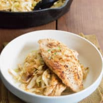 Resting on a burlpa npakin atop a wood slat tabletop is Seared chicken breast on top of a bed of smoky cabbage noodles in a white bowl. In the background sit the cast iron pan with chicken and cabbage noodles.