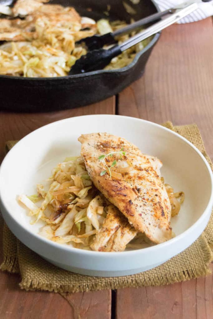 Resting on a burlpa npakin atop a wood slat tabletop is Seared chicken breast on top of a bed of smoky cabbage noodles in a white bowl. In the background sit the cast iron pan with chicken and cabbage noodles.