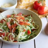On a white slat table sits a beige pottery bowl with a colourful cucumber, tomato, onion salad mixed in it. A bowl of parmesan cheese, two whole tomatoes, and zesty italian dressing in a glass dressing dispenser sit nearby.