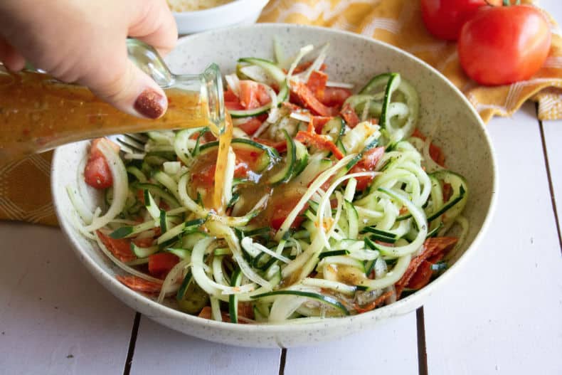 Ingredients for the zesty italian cucumber salad are added to a creamy white pottery bowl while a glass salad dressing bottle is held above and drizzles the italian dressing onto the salad.