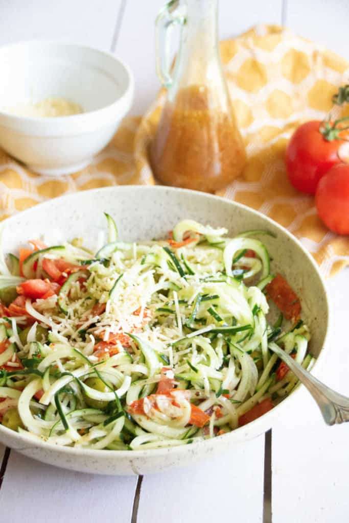 Image of zesty Italian cucumber salad in a speckled bowl on a wood slat table top. Bottle of Italian dressing is beside the bowl, a yellow tea towel, Parmesan and whole tomatoes are in the background