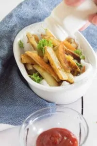 Homemade French Fries sit in a white ceramic bowl, on a blue tea towel on top of a white wood table top. A glass bowl of ketchup sits next to it. A white salt shaker, shakes salt above it.