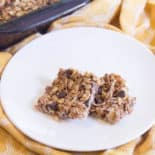 Two square choloate chip granola bars sit on a round white plate on top of a yellow dish towel.