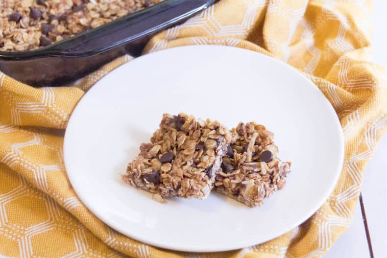 Two square chocolate chip granola bars sit on a round white plate on top of a yellow dish towel.
