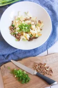 Apple, green onions, nuts, and parsley are tossed together in a medium-sized bowl on a blue tea towel. A wooden cutting board with chopped nuts and green onion sits by the bowl.