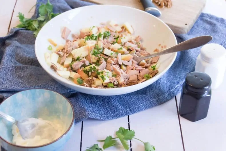 Tuna is added to the nut, apple, onion mixture in a white bowl. Dressing sits nearby in a small blue bowl. Salt and pepper shakers sit to the right of the salad bowl.