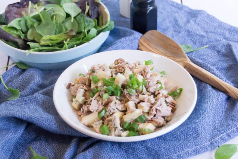 Waldorf Tuna Salad in a white bowl, on top of a blue tea towel. A bowl of baby spinach sits in the background, a wooden spoon sits nearby.