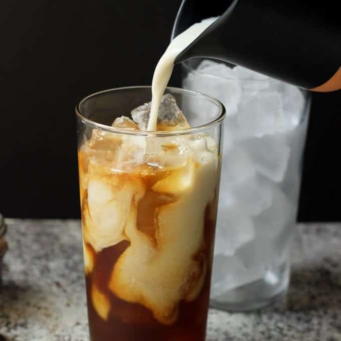 milk pouring into a glass of iced coffee with a container of ice behind it