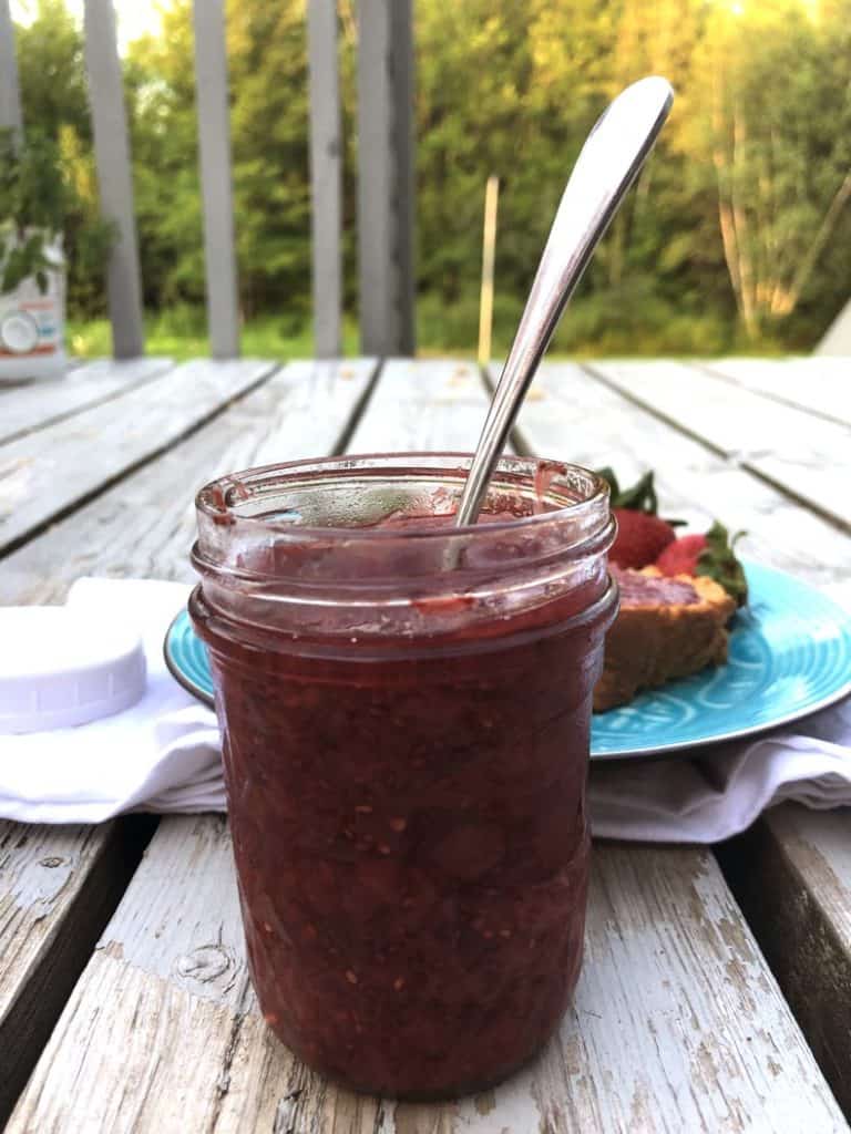 a jar of homemade strawberry jam with a blurred out background of trees, plus a plate of jam and bread