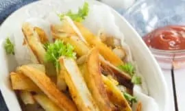 Homemade French Fries sit in a white ceramic bowl, on a blue tea towel on top of a white wood table top. A glass bowl of ketchup sits next to it. A white salt shaker, shakes salt above it.