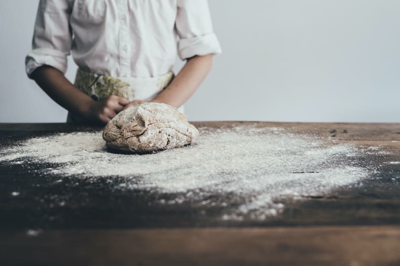 Image is of a kitchen work surface,  heavily dusted with flour, a ball of dough sits on the floured surface, and the torso of a chef is in the background, behind the dough.