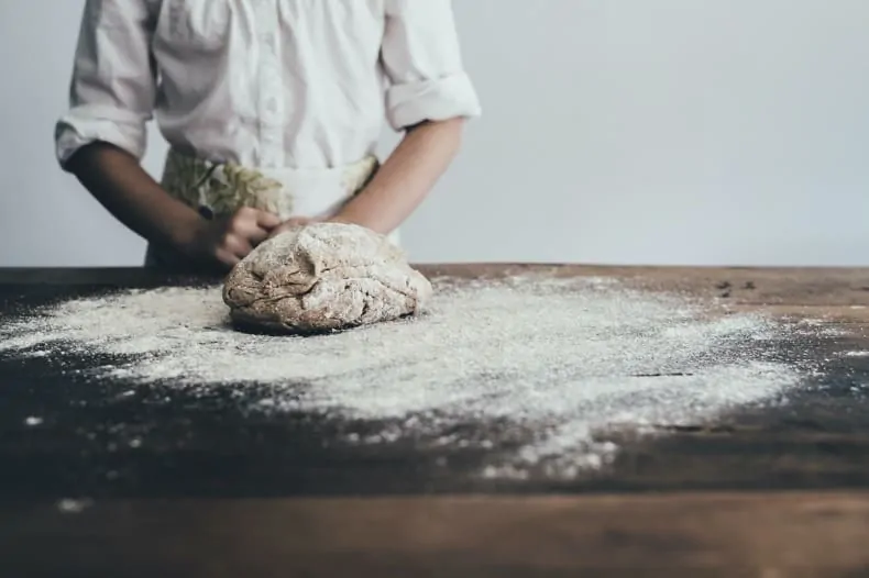 Image is of a kitchen work surface, heavily dusted with flour, a ball of dough sits on the floured surface, and the torso of a chef is in the background, behind the dough.