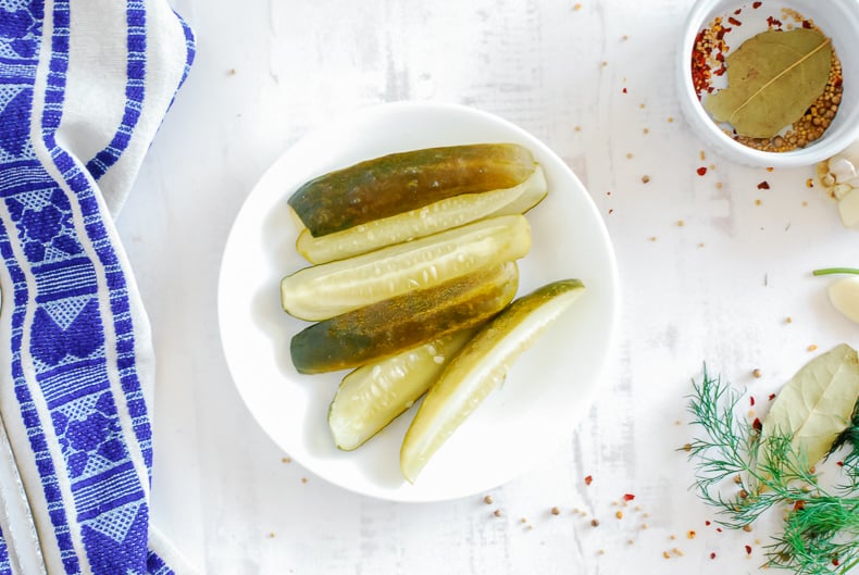 Pickles cut lengthwise into spears on a white plate, pickling ingredients, and a tea towel sit nearby.