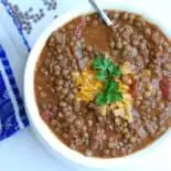 Chili lentils in a white bowl on a white surface with a blue patterned dishtowel. Garnished with cheese and a sprig of green.