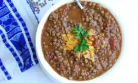 Chili lentils in a white bowl on a white surface with a blue patterned dishtowel. Garnished with cheese and a sprig of green.