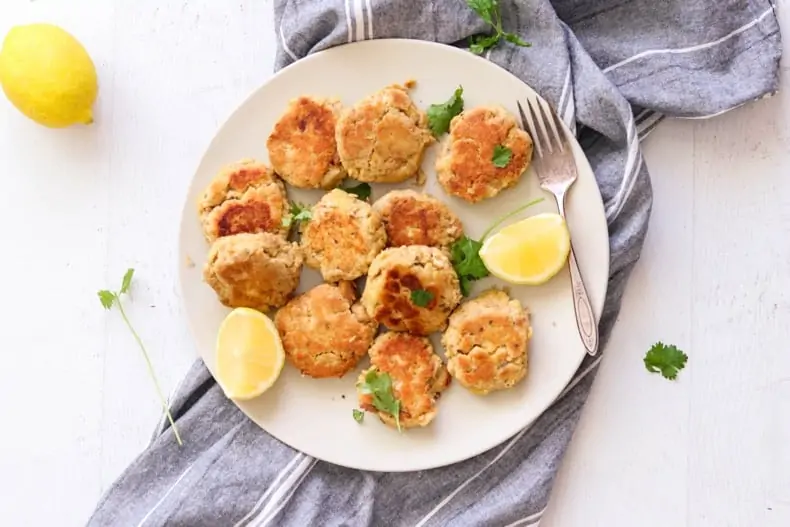 Gluten-free salmon cakes on a white plate and white surface, with a grey striped dishtowel, and some parsley and lemon garnish.