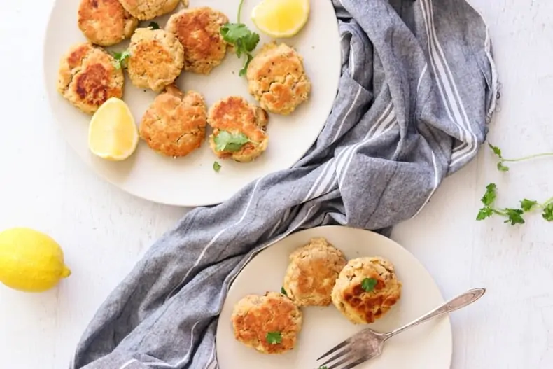 Two plates of gluten-free salmon patties on a white surface with a grey tea towel and lemons as garnish