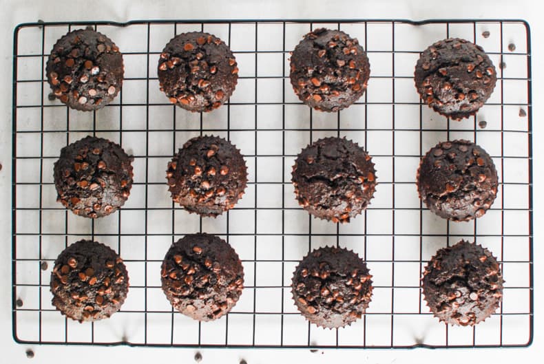 Overhead shot of a dozen coconut flour chocolate muffins on a black cooling rack and white surface,