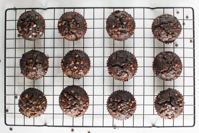 Overhead shot of a dozen coconut flour chocolate muffins on a black cooling rack and white surface,