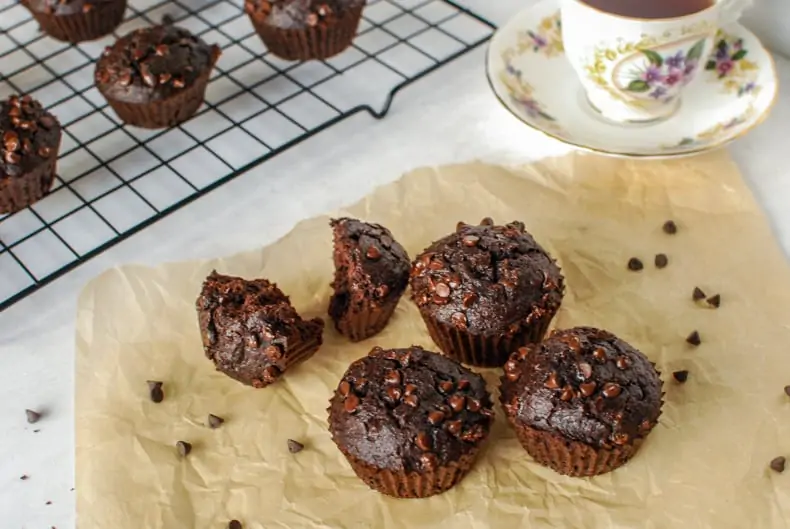 45 degree shot of some coconut flour chocolate muffins on a piece of parchment paper with a cup of tea and muffins on a cooling rack in the background