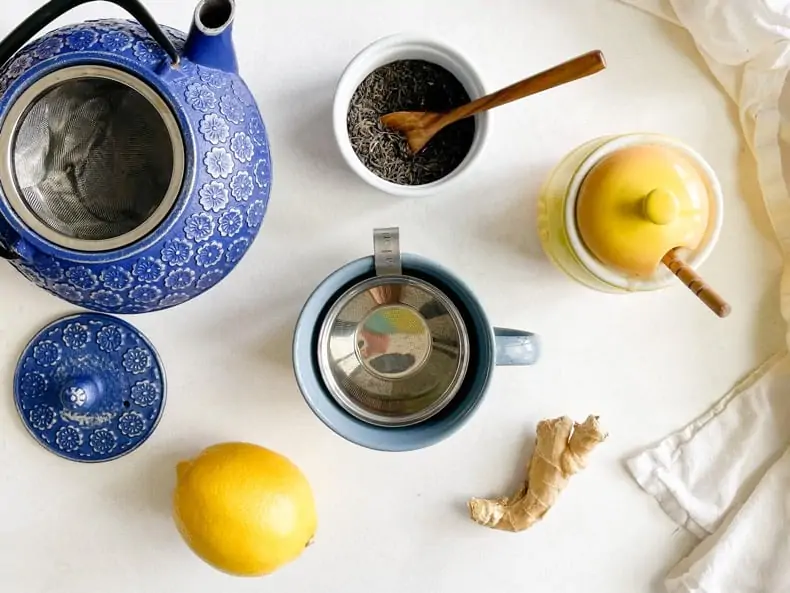 Ingredients for stomach debloating tea remedy on a white surface, including a mug in the center, fresh ginger, fresh lemon, honey, loose green tea, and a blue cast iron teapot