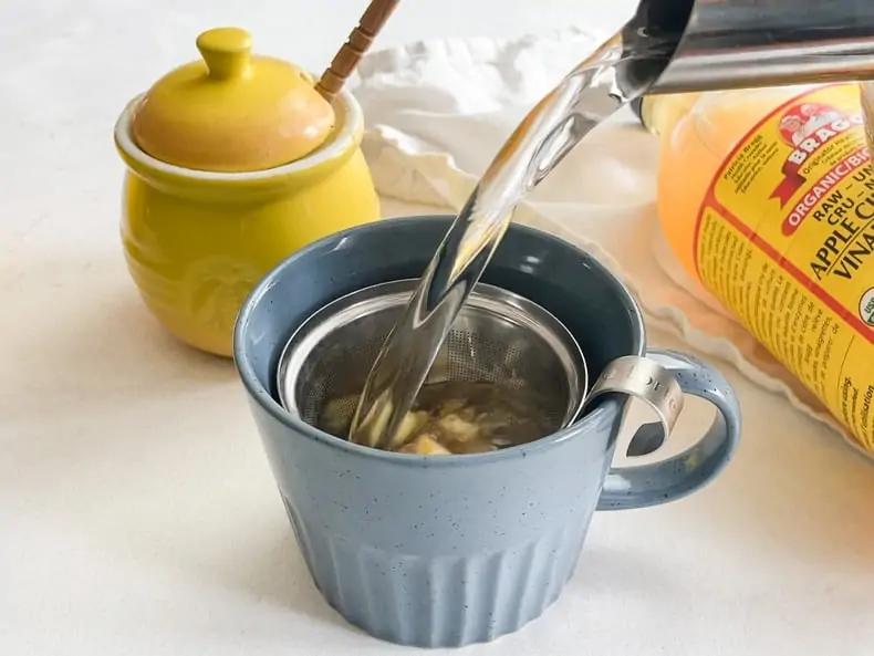 Pouring water from the kettle into a blue mug with minced fresh ginger to make debloating tea remedy