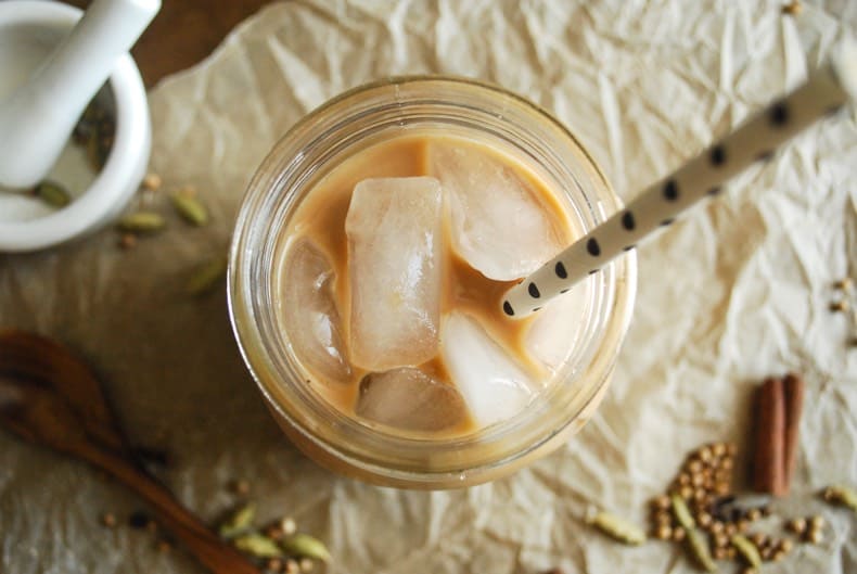 An overhead shot of the iced chai latte with ice cubes, with a crinkled parchment paper surface, some scattered spices, and a mortar and pestle in the background.