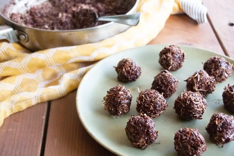A plate of chocolate coconut macaroons sitting on a surface looking tempting and delicious, with a pot and napkin in the background.