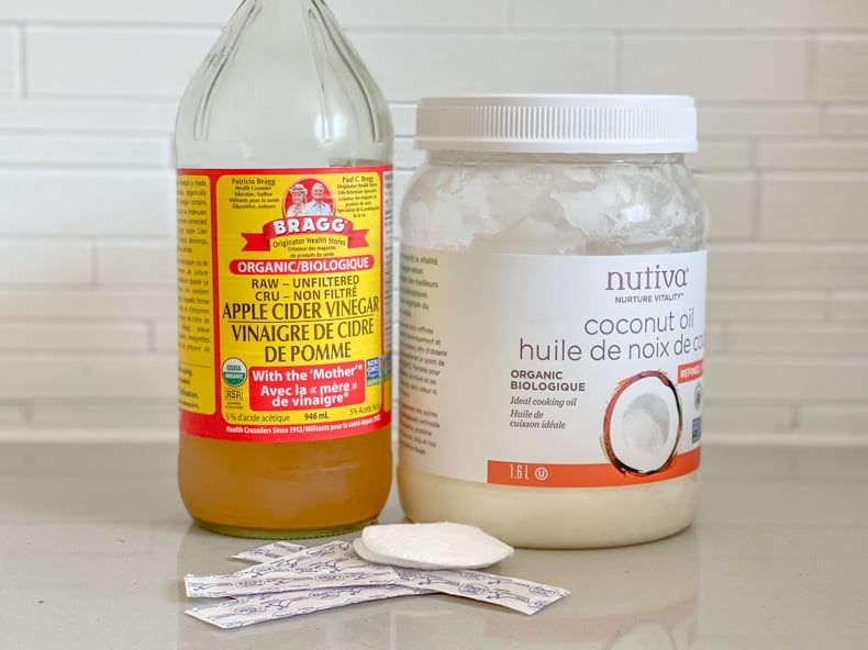 A close of of a bottle of apple cider vinegar and a jar of coconut oil sitting on a counter with a white background, with bandaids and cotton pads beside them.