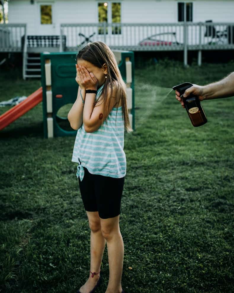 a young girl standing in a backyard with green grass, covering her face while being sprayed with homemade mosquito repellent in a brown glass spray bottle.